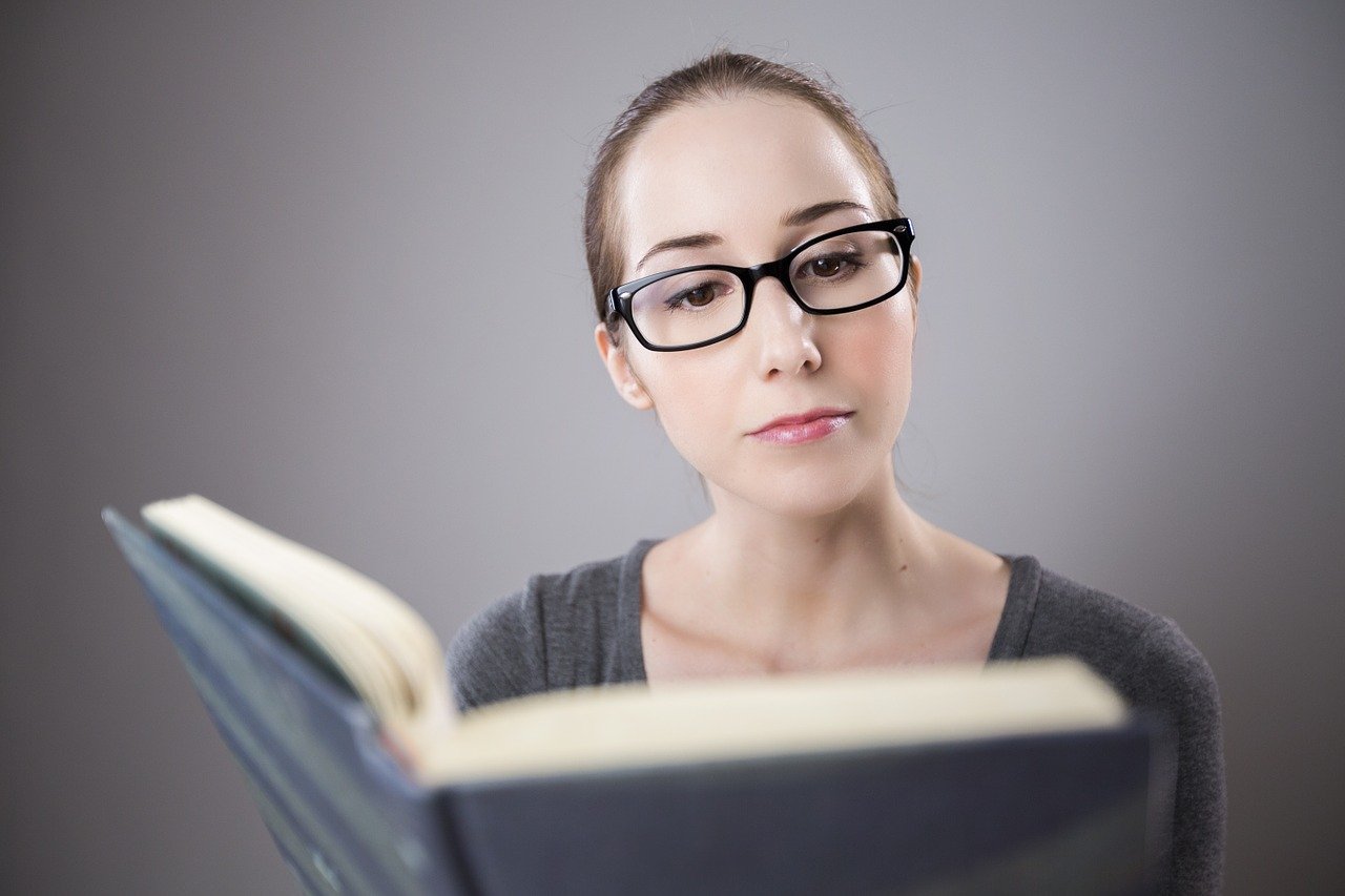 Uma mulher jovem de óculos lendo um livro. Imagem de PourquoiPas por Pixabay