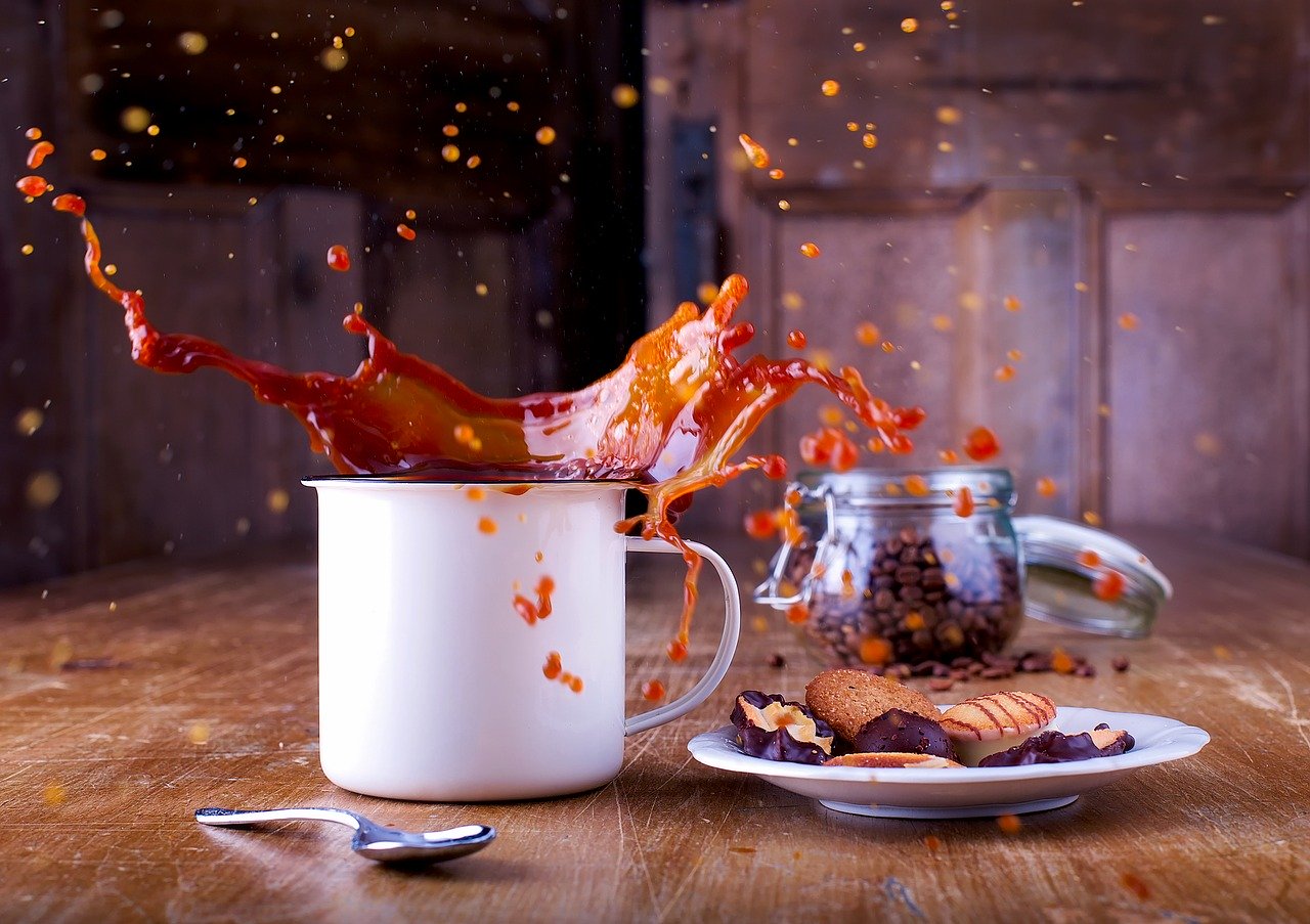 Uma xícara de café espalhado por toda mesa de madeira. Ao lado, um pires com biscoitos de cholocate, uma colher de sobremesa e ao fundo a cafeteira. Imagem de David Mark por Pixabay